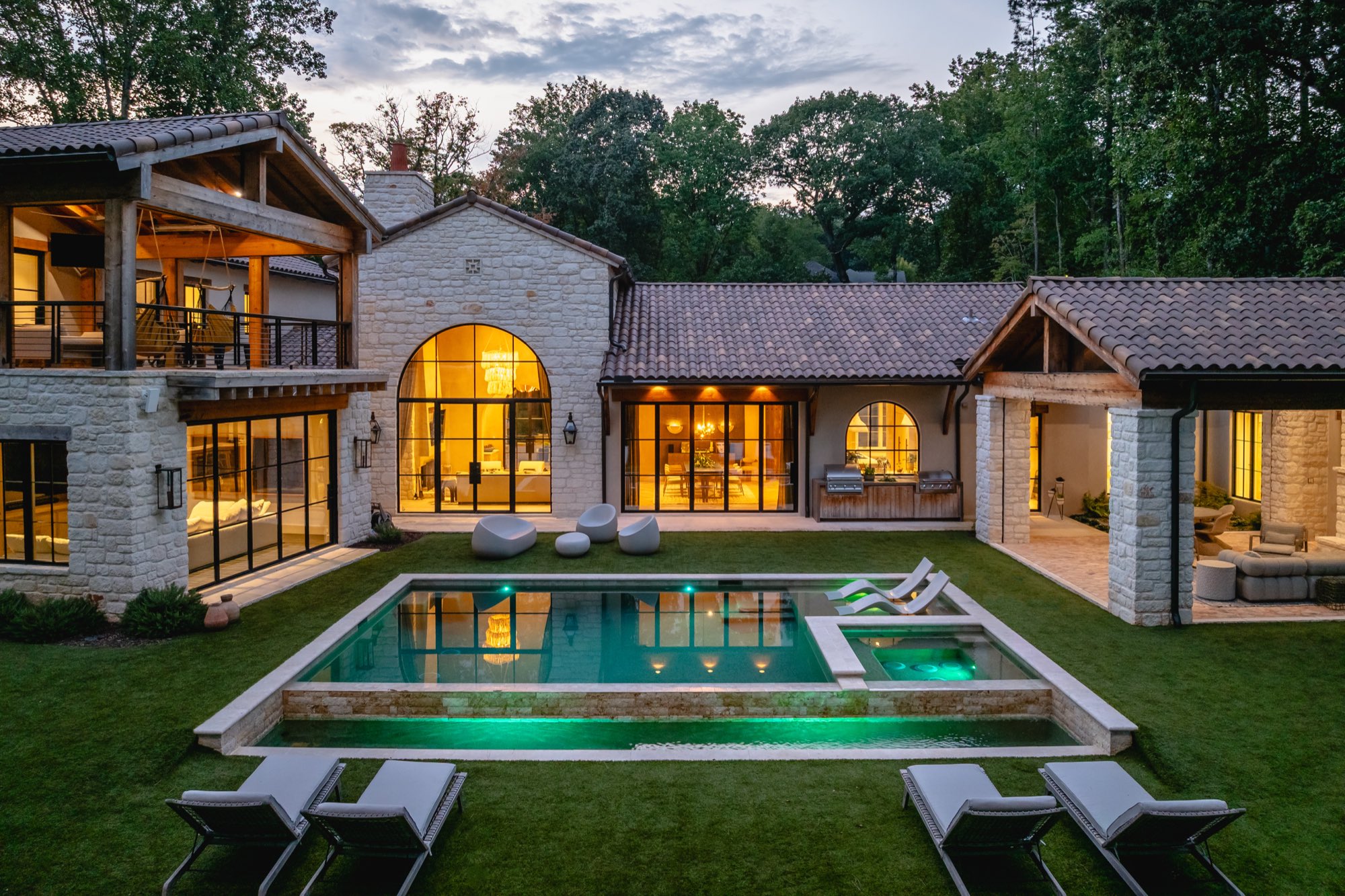 A stone and timber home at dusk, arched windows glowing over the courtyard pool