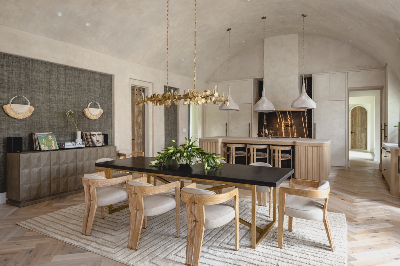 A dining table under a barrel-vaulted ceiling, the kitchen glowing beyond