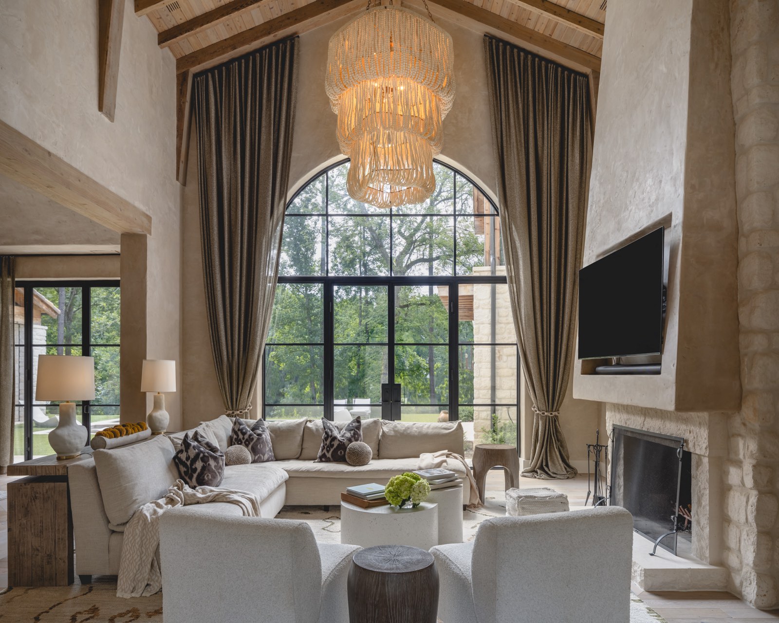 A vaulted great room with timber beams, a plaster fireplace, and a tall arched steel window over deep white sofas