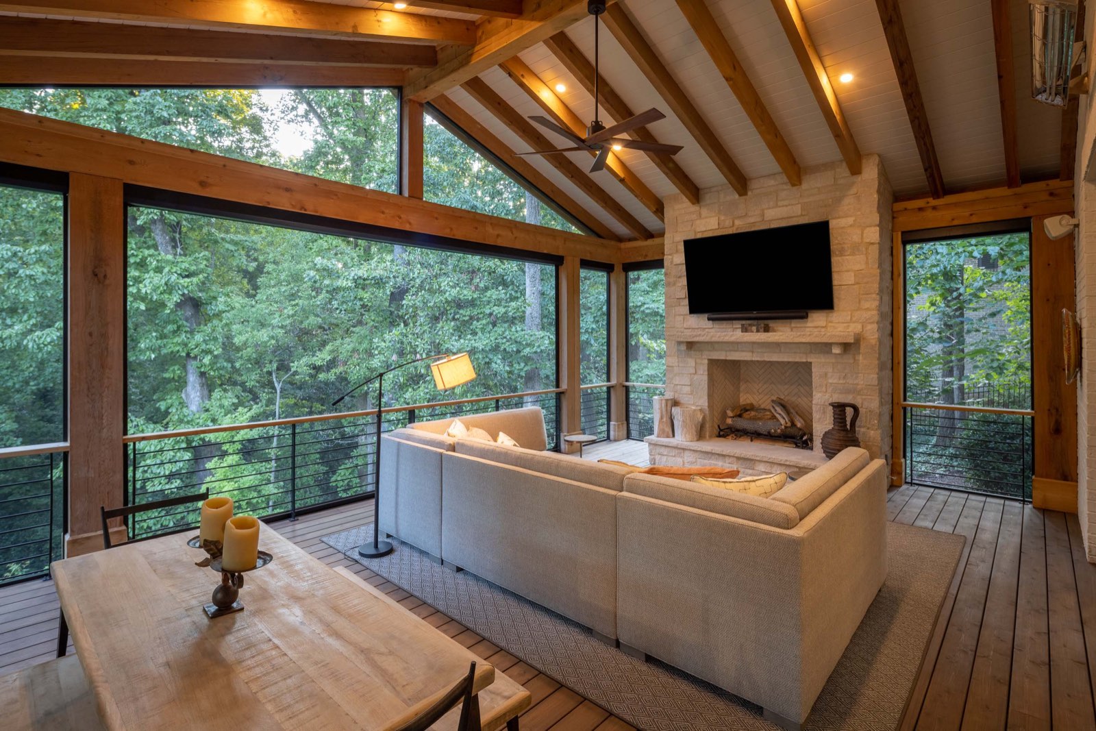 A screened porch with timber trusses and a stone fireplace, deep sofas facing the trees